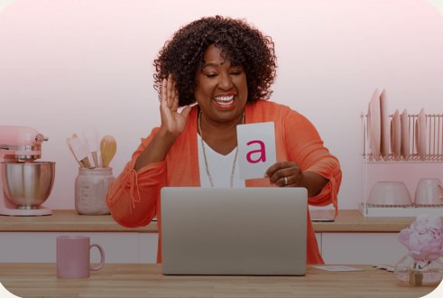 A smiling adult waves at a laptop while holding up a flashcard with the letter a