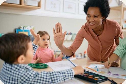 A teacher high-fives a young child at a classroom table while another child looks on