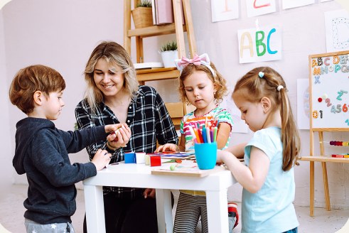 An adult helps three children with a craft activity at a small white table
